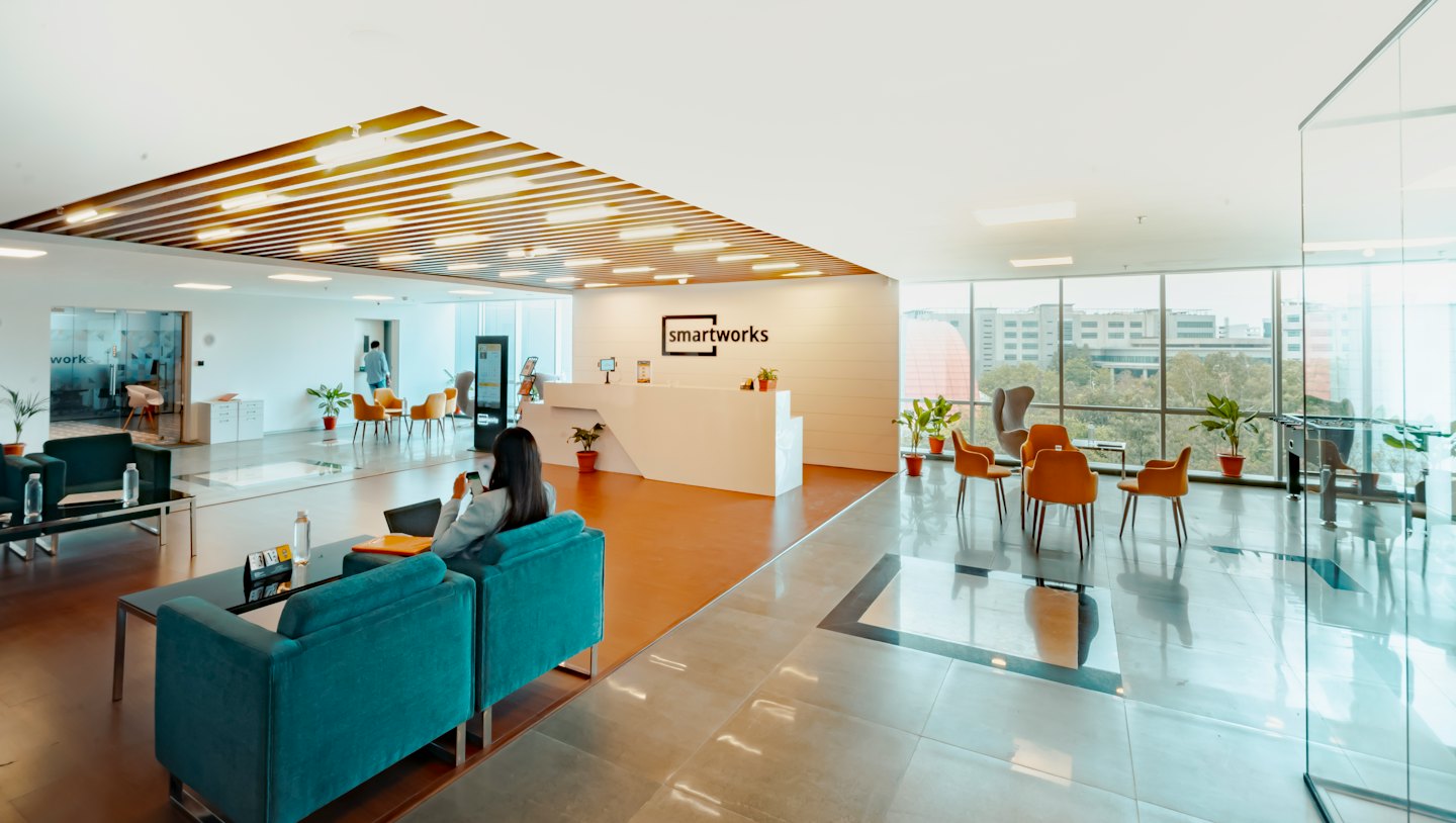 Woman on teal sofa uses phone in modern office reception.